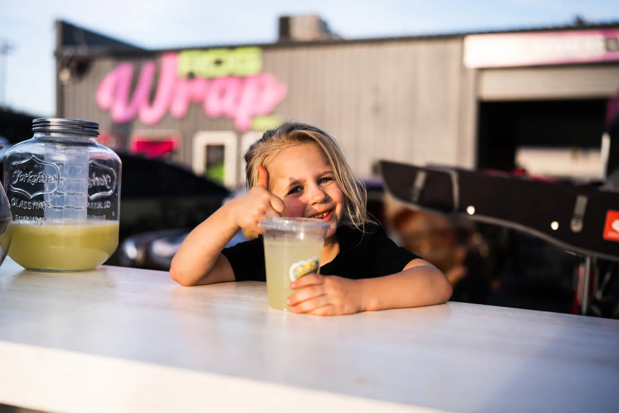 A young girl gives a thumbs up from the lemonade stand at the Kool April Nights block party, the neon RDG Wrap sign behind her